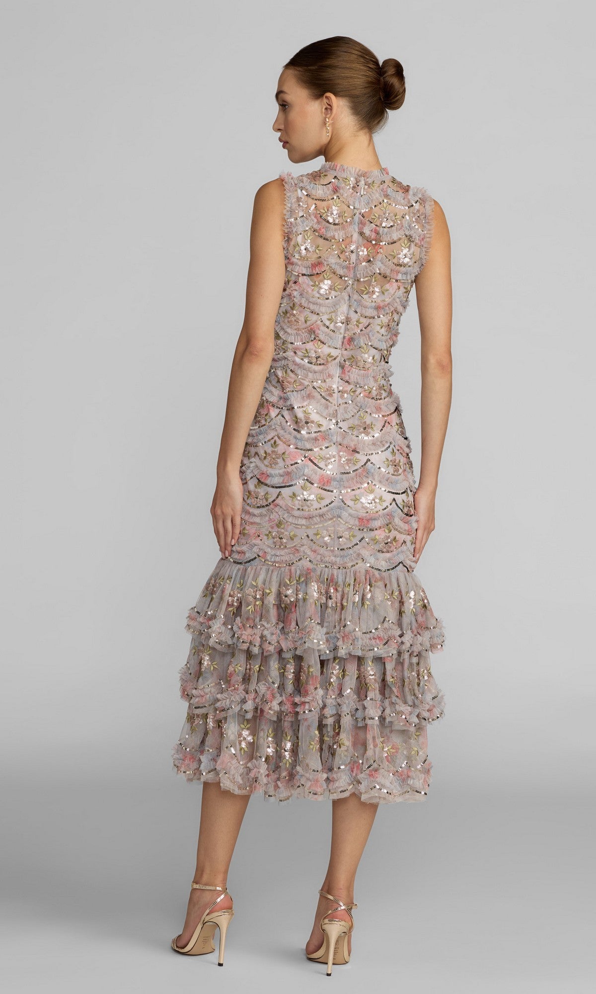 A woman stands with her back to the camera, her hair elegantly styled in a bun, wearing the Midi Party Dress: Mac Duggal 8412โa sleeveless floral midi dress adorned with knee-length ruffled layers, delicate metallic accents, and intricate embroidery. She finishes the look with high heels for a sophisticated party ensemble inspired by Mac Duggalโs signature elegance.